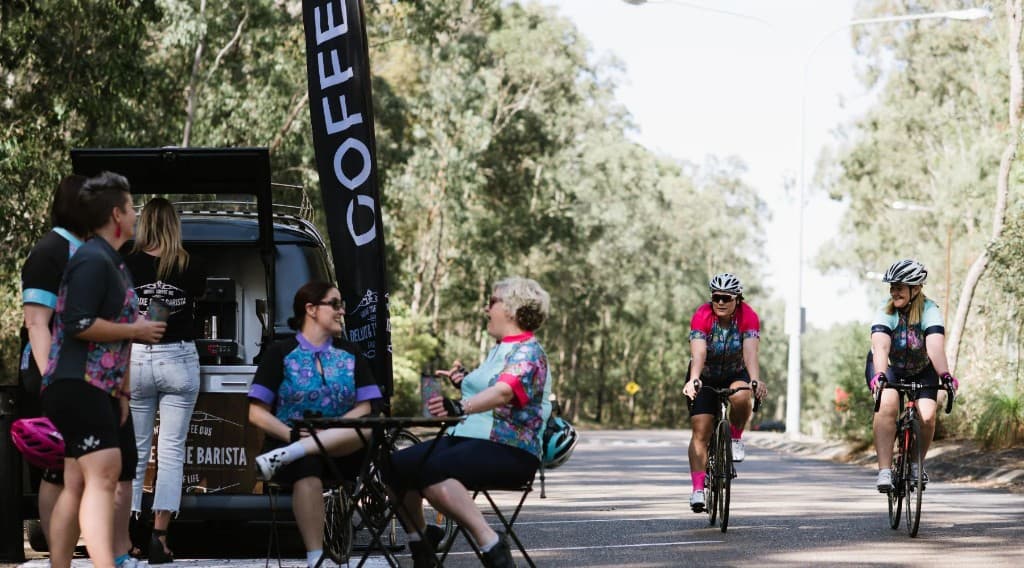 Cyclists taking a break at a mobile coffee van beside a tree-lined road